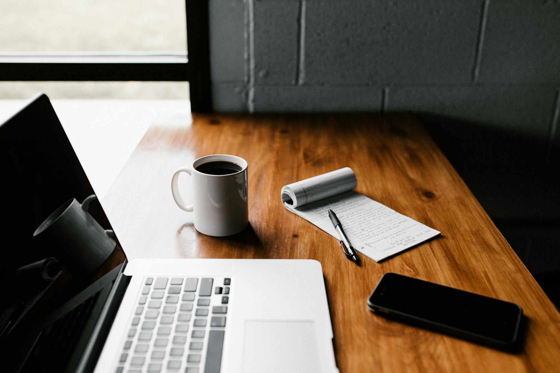 Laptop on a desk beside a cup of coffee, a notepad, and a phone.